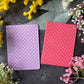 Two rectangular cards or envelopes with embossed diamond patterns, one light purple and one coral pink, arranged on a dark textured surface surrounded by yellow and pink flowers.