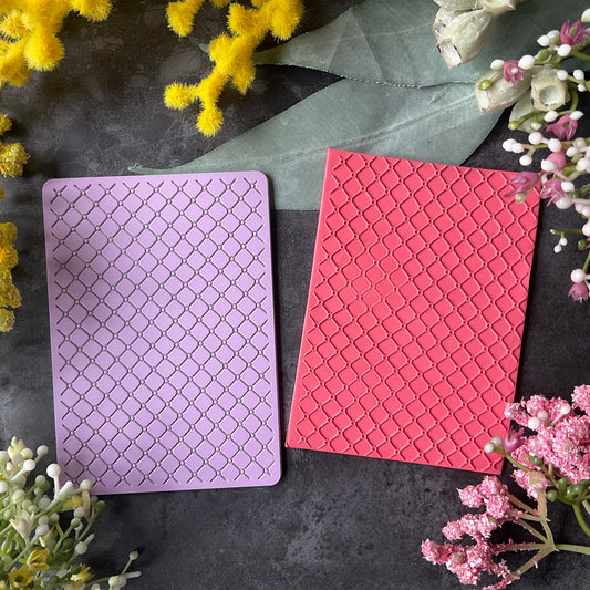 Two rectangular cards or envelopes with embossed diamond patterns, one light purple and one coral pink, arranged on a dark textured surface surrounded by yellow and pink flowers.