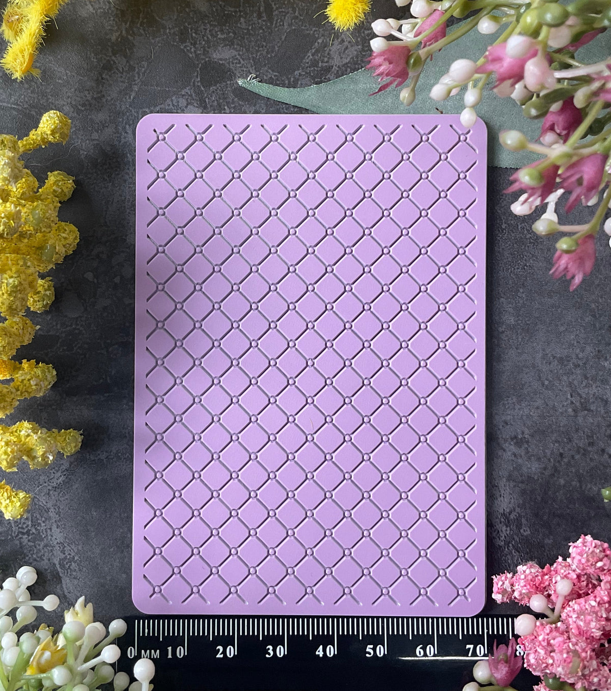 A rectangular lavender purple silicone stamp with a raised trellis pattern, displayed on a dark gray textured background and surrounded by yellow and pink flowers, with a ruler at the bottom indicating its 7cm width.