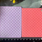 Two rectangular embossed stamp or plaque blocks, one lavender and one coral pink, displayed side-by-side on a dark textured surface with a black ruler at the bottom for scale and flowers around the edges.