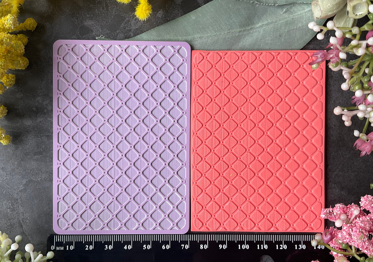 Two rectangular embossed stamp or plaque blocks, one lavender and one coral pink, displayed side-by-side on a dark textured surface with a black ruler at the bottom for scale and flowers around the edges.