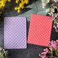 Two rectangular embossed cardstock sheets with a repeating trellis pattern, one in light lavender and one in coral pink, styled flat-lay on a dark textured surface with yellow and pink flowers.