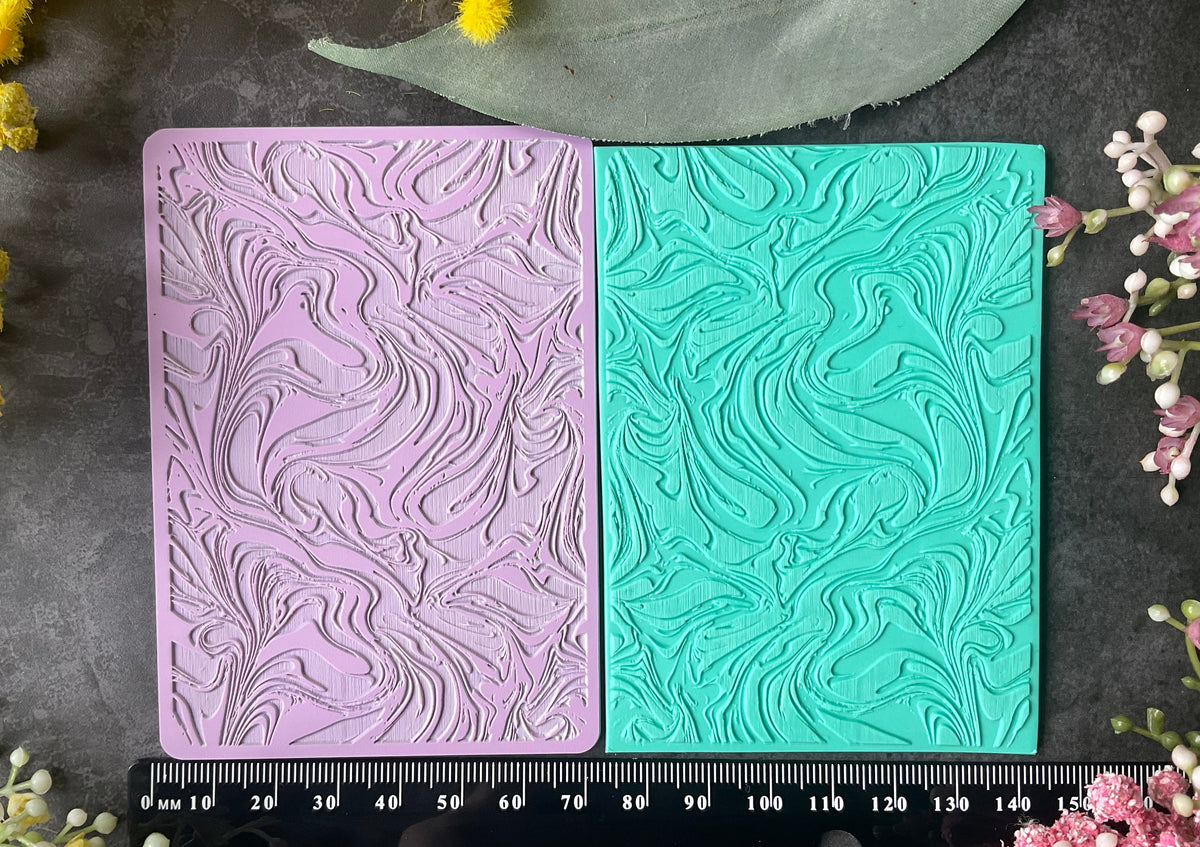 Two rectangular embossed stamp blocks or tiles shown side by side on a dark textured surface, one in light lavender and one in mint green, with a ruler along the bottom for scale and flowers around the edges.