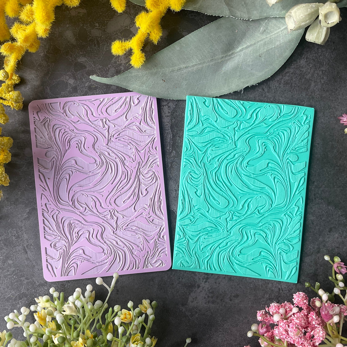 Two rectangular embossed stamp or cardstock pieces in pastel purple and turquoise, arranged on a dark textured surface with yellow and pink flowers around them.