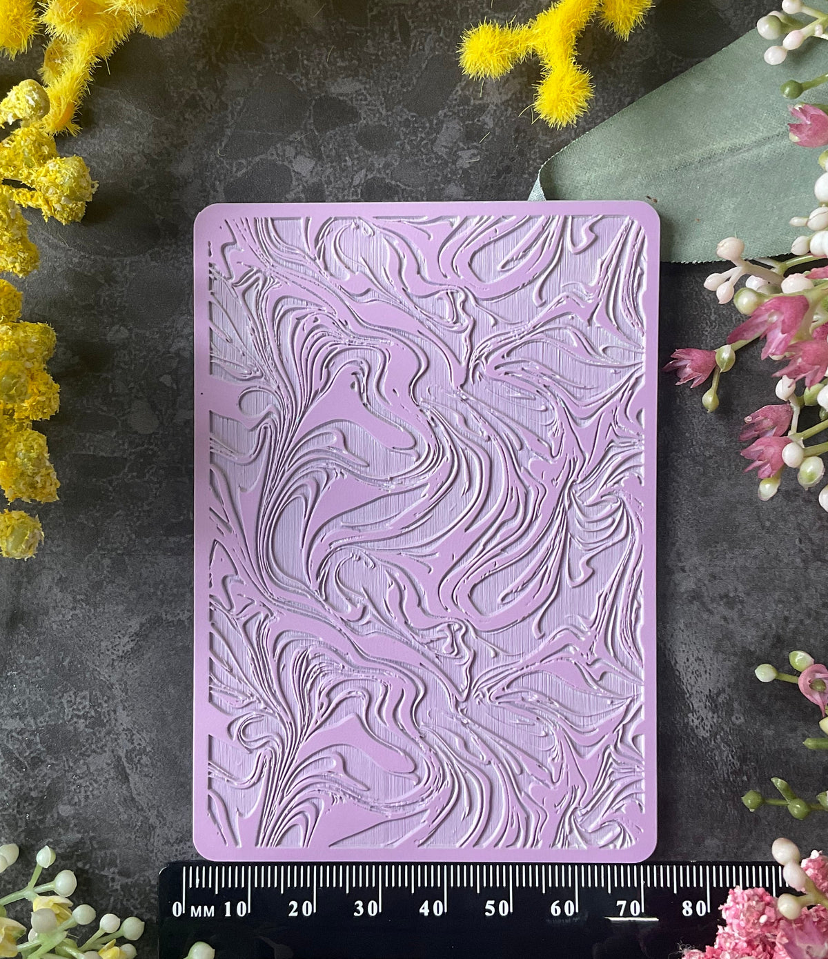 A lavender rectangular embossed stamp or mold with a swirling floral pattern, displayed on a dark gray surface with a black measuring ruler underneath and yellow and pink flowers framing the scene.
