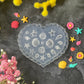 A heart-shaped translucent silicone mold decorated with embossed seashells and starfish, surrounded by colorful miniature seashells and starfish decorations on a dark textured surface with yellow and pink flowers.