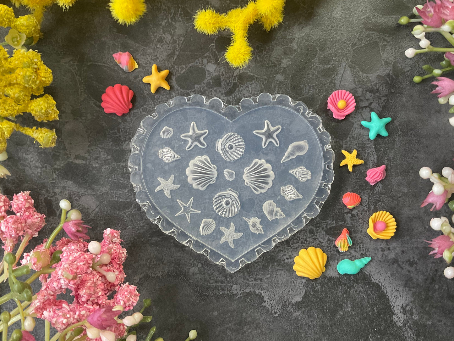A heart-shaped translucent silicone mold decorated with embossed seashells and starfish, surrounded by colorful miniature seashells and starfish decorations on a dark textured surface with yellow and pink flowers.