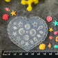 A heart-shaped translucent plastic mold with embossed seashell and starfish patterns, displayed on a dark textured background with a black ruler at the bottom for scale, surrounded by yellow and pink flowers.
