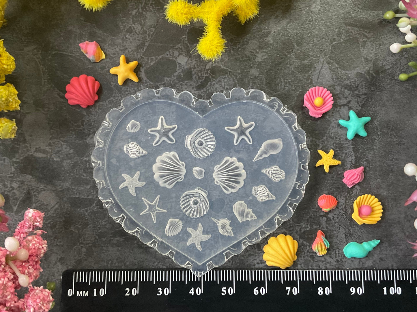 A heart-shaped translucent plastic mold with embossed seashell and starfish patterns, displayed on a dark textured background with a black ruler at the bottom for scale, surrounded by yellow and pink flowers.