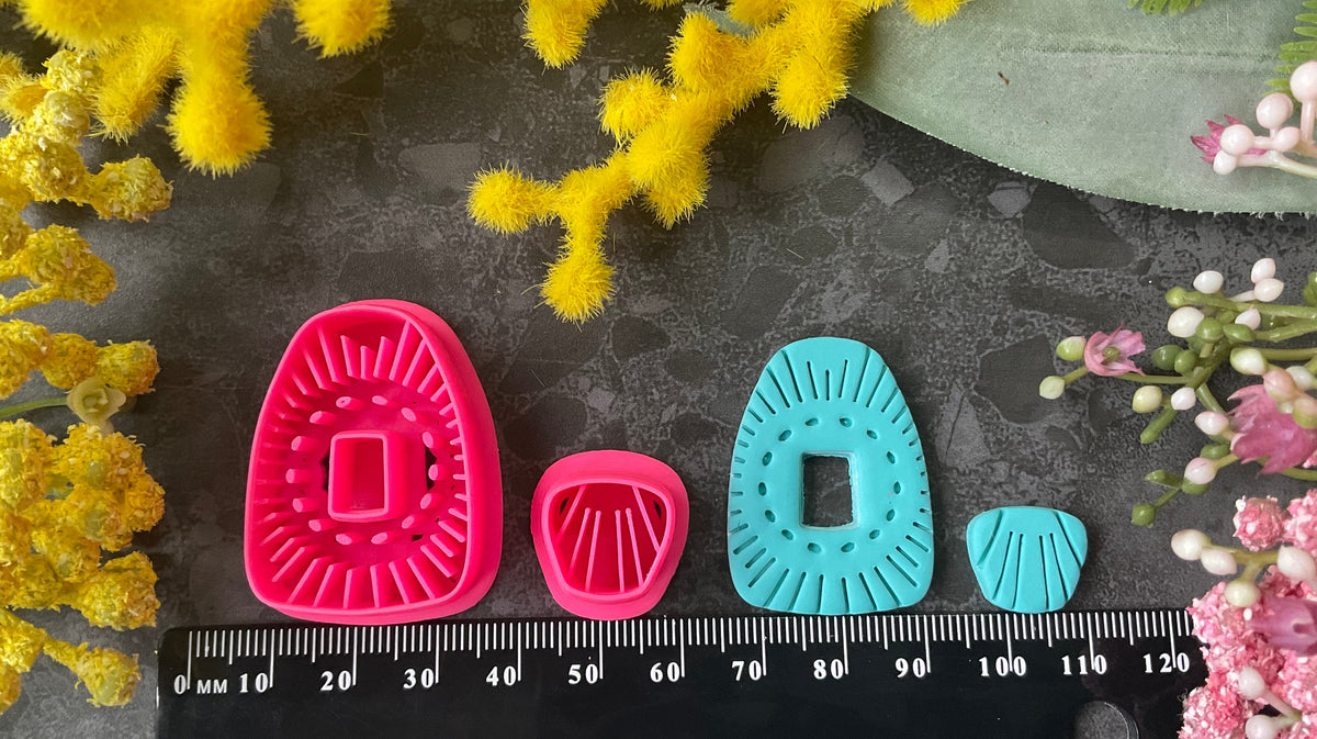 Bright pink 3d printed polymer clay cutters in bright pink and turquoise arranged on a dark textured surface, surrounded by yellow and pink flowers. Solstice Bloom Polymer Clay Cutter Set