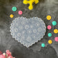 A heart-shaped clear plastic stamp mold with embossed floral patterns, displayed on a dark gray textured surface surrounded by yellow and pink flowers and small colorful flower stamps.