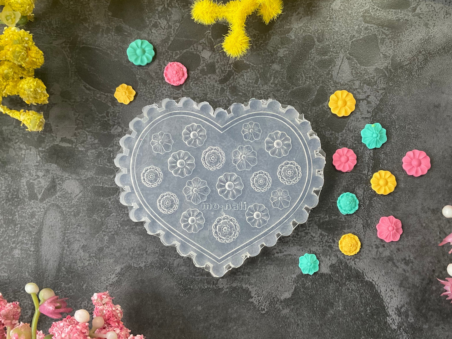 A heart-shaped clear plastic stamp mold with embossed floral patterns, displayed on a dark gray textured surface surrounded by yellow and pink flowers and small colorful flower stamps.