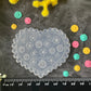 A heart-shaped clear plastic stamp mold with floral patterns, displayed on a dark textured surface with a black ruler at the bottom for scale, surrounded by small colorful flower petals and yellow flowers.