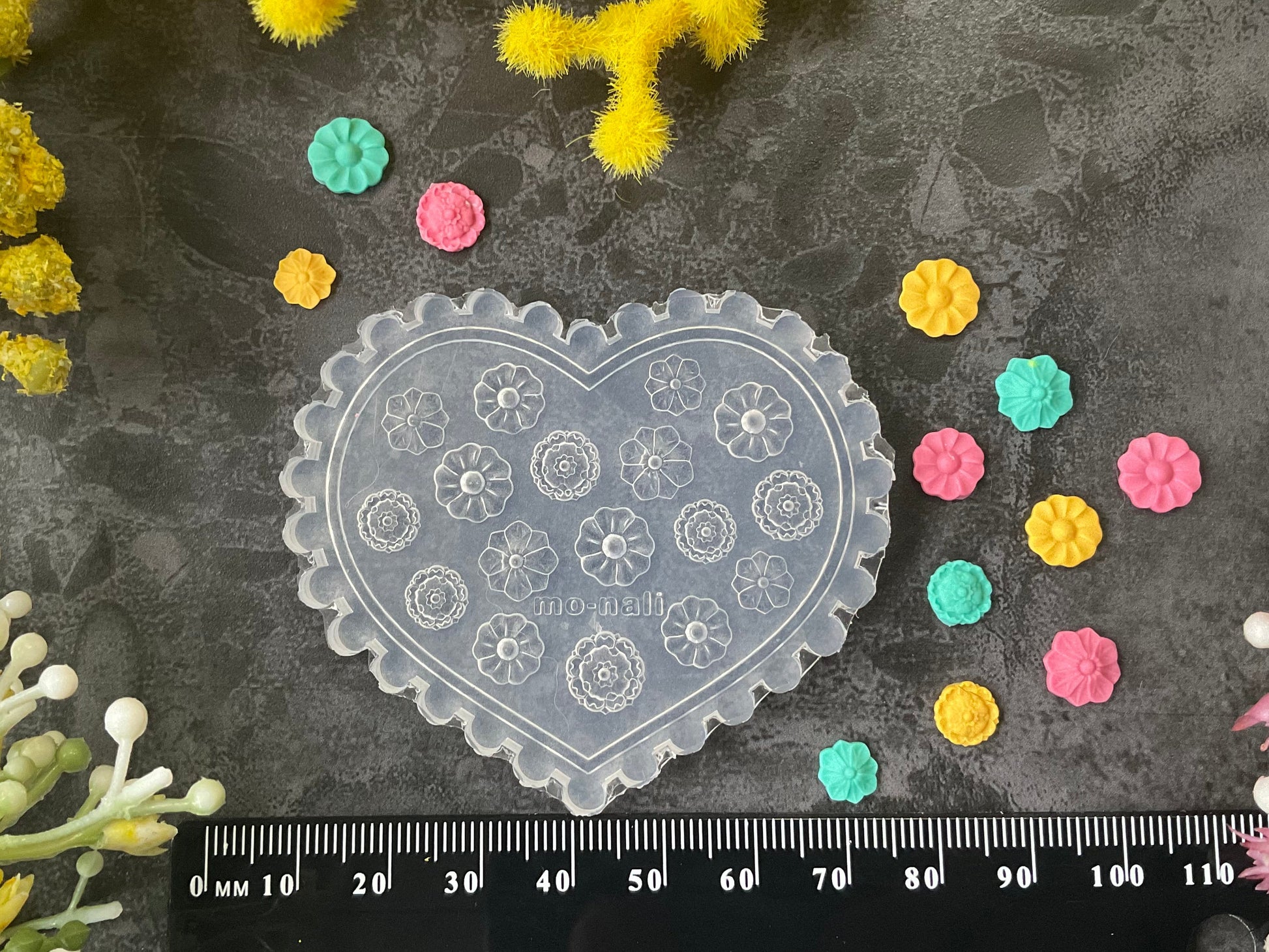 A heart-shaped clear plastic stamp mold with floral patterns, displayed on a dark textured surface with a black ruler at the bottom for scale, surrounded by small colorful flower petals and yellow flowers.