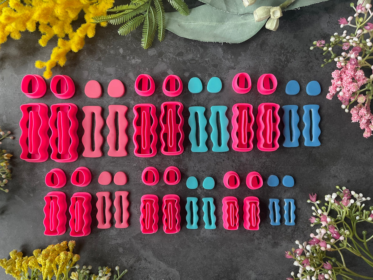 A set of colorful 3d printed polymer clay cutters arranged in rows on a dark textured surface, surrounded by decorative flowers. The cutters come in three main colors: bright pink, coral pink, and teal blue, with various shapes including circles, ovals, and wavy patterns. Squiggly Ripple Pillar Polymer Clay Cutter Set