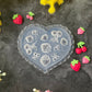 A heart-shaped clear silicone mold with embossed strawberry and floral designs, surrounded by small pink and red strawberry charms on a dark textured surface with yellow and pink flowers.