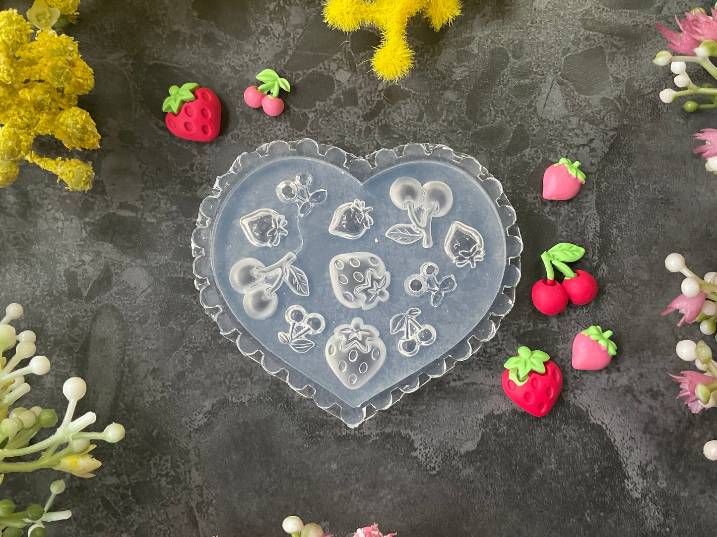 A heart-shaped clear silicone mold with embossed strawberry and floral designs, surrounded by small pink and red strawberry charms on a dark textured surface with yellow and pink flowers.