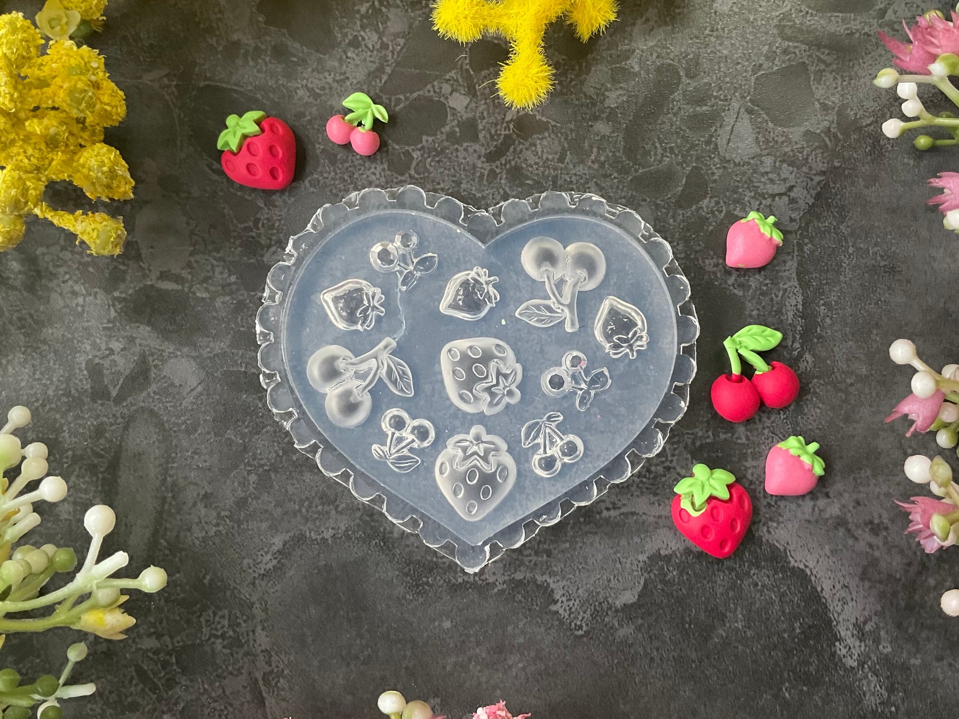 A heart-shaped clear silicone mold with embossed strawberry and floral designs, surrounded by small pink and red strawberry charms on a dark textured surface with yellow and pink flowers.
