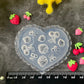 A heart-shaped translucent plastic mold with embossed strawberry and floral designs, displayed on a dark textured surface with a black ruler at the bottom and small strawberry and berry decorations arranged around it.