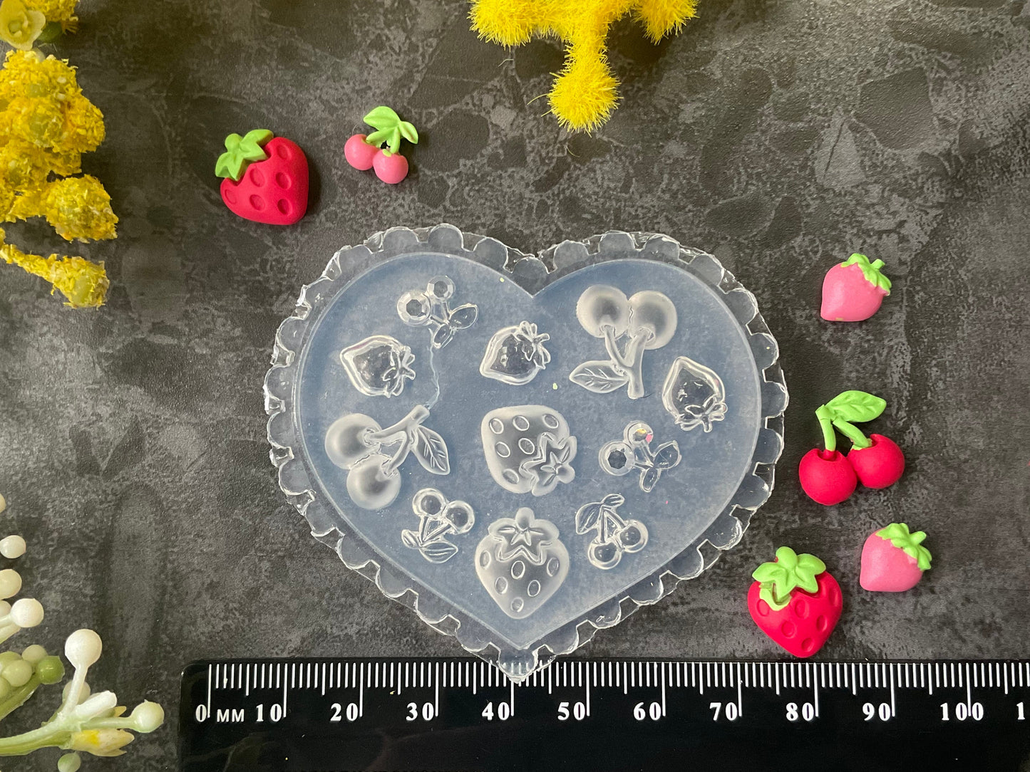 A heart-shaped translucent plastic mold with embossed strawberry and floral designs, displayed on a dark textured surface with a black ruler at the bottom and small strawberry and berry decorations arranged around it.