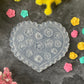 A heart-shaped clear plastic craft mold with embossed floral and geometric patterns, displayed on a dark textured surface surrounded by small colorful flower-shaped embellishments and yellow decorative stems.