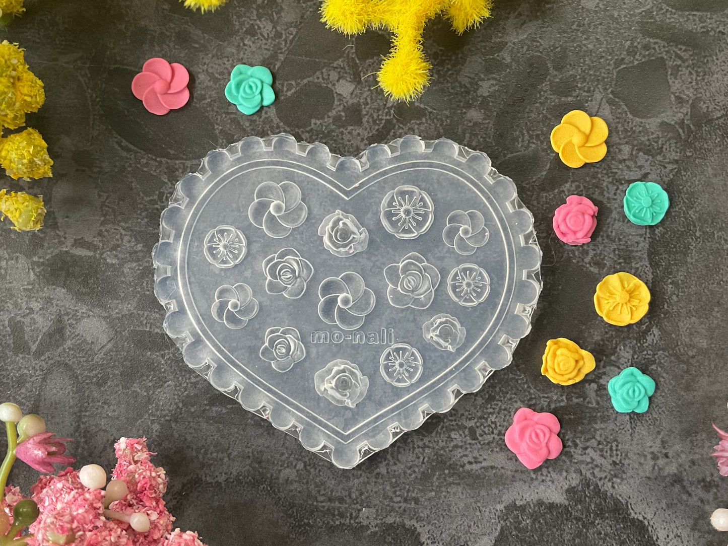 A heart-shaped clear plastic craft mold with embossed floral and geometric patterns, displayed on a dark textured surface surrounded by small colorful flower-shaped embellishments and yellow decorative stems.