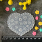 A heart-shaped clear plastic craft mold with embossed floral designs, displayed on a dark textured surface with a black ruler at the bottom and small colorful flower-shaped pieces scattered around. Yellow and pink flowers frame the top and sides of the scene.