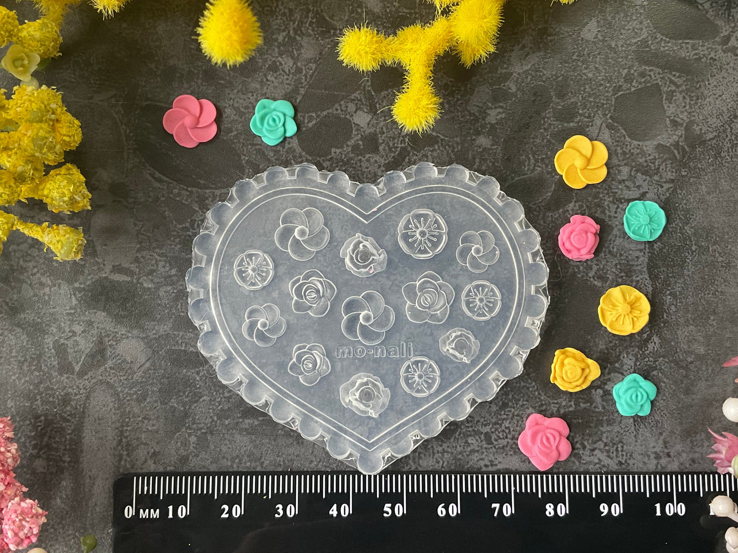 A heart-shaped clear plastic craft mold with embossed floral designs, displayed on a dark textured surface with a black ruler at the bottom and small colorful flower-shaped pieces scattered around. Yellow and pink flowers frame the top and sides of the scene.