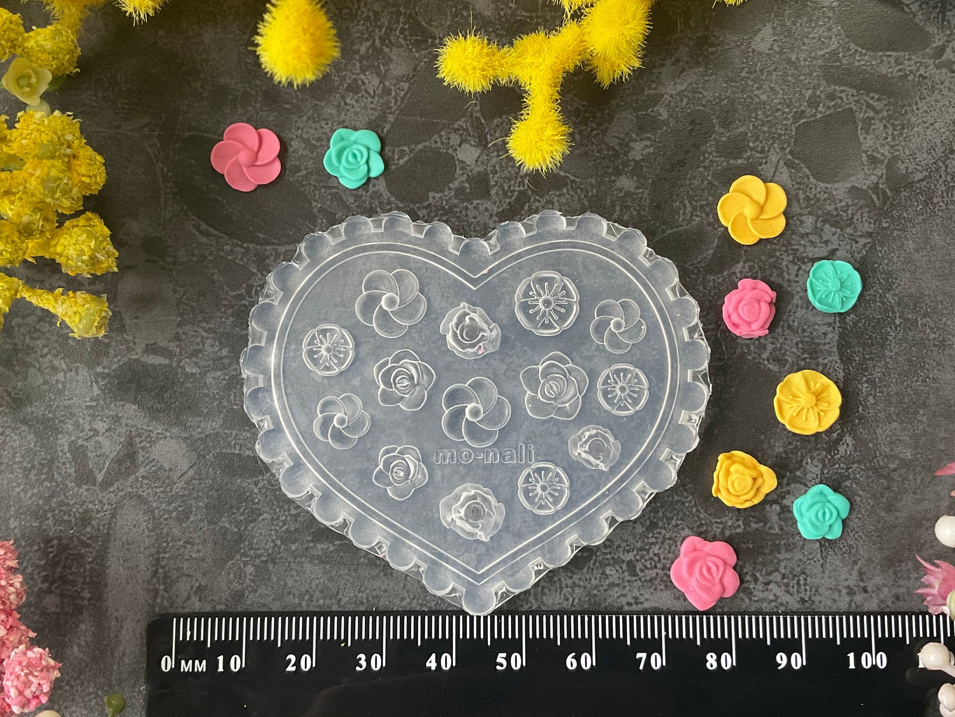 A heart-shaped clear plastic craft mold with embossed floral designs, displayed on a dark textured surface with a black ruler at the bottom and small colorful flower-shaped pieces scattered around. Yellow and pink flowers frame the top and sides of the scene.