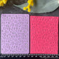 Two rectangular embossed stamping plates, one light purple and one bright pink, displayed side-by-side on a dark gray textured surface with a black ruler below for scale and decorative flowers around the edges.
