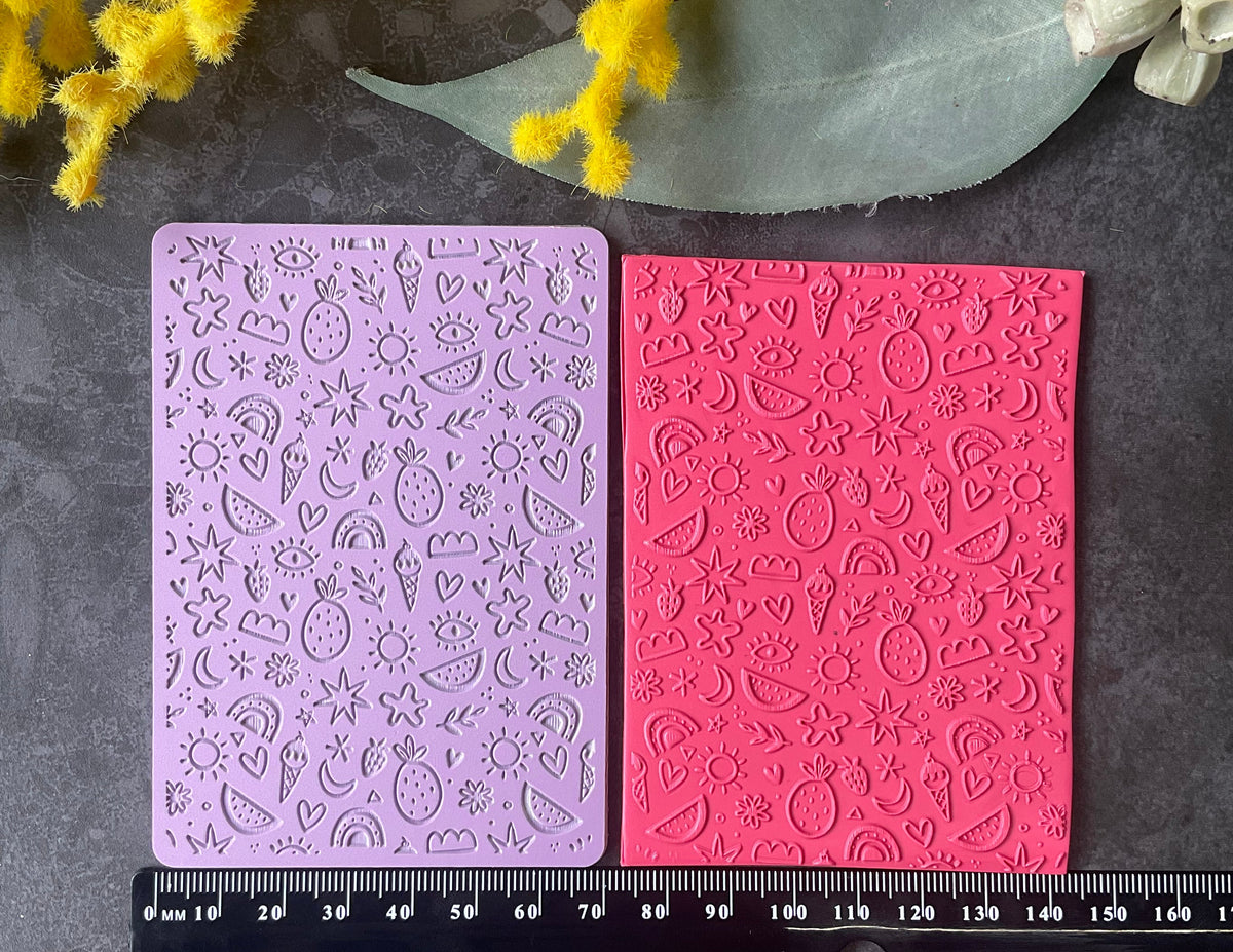 Two rectangular embossed stamping plates, one light purple and one bright pink, displayed side-by-side on a dark gray textured surface with a black ruler below for scale and decorative flowers around the edges.