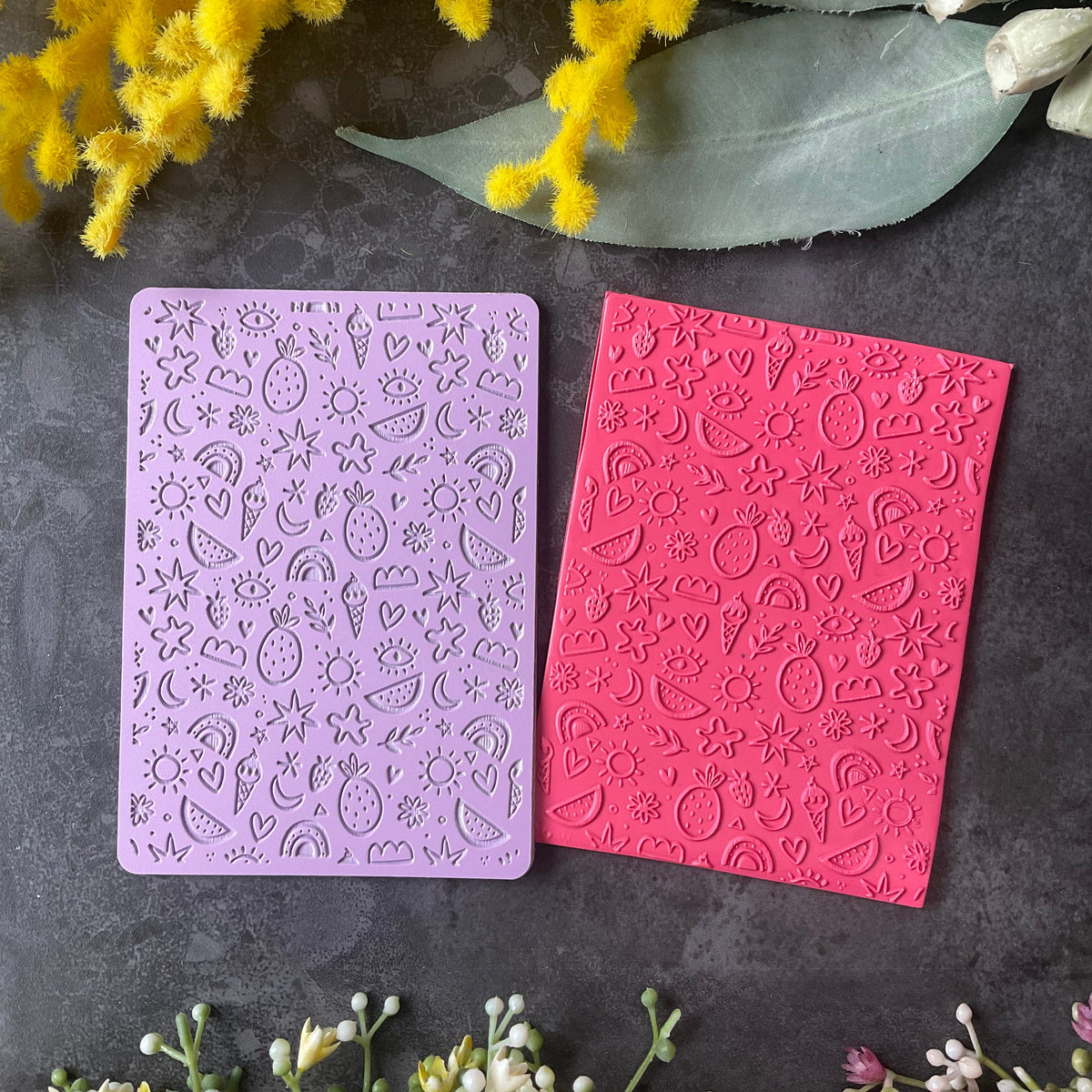Two rectangular embossed stamping plates, one lavender purple and one bright pink, displayed on a dark gray textured surface with decorative yellow and white flowers.