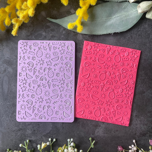 Two rectangular embossed stamping plates, one lavender purple and one bright pink, displayed on a dark gray textured surface with decorative yellow and white flowers.