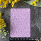 A lavender purple silicone stamp mold with embossed summer-themed designs like ice cream, suns and rainbows, and flowers, displayed on a dark gray textured surface with a black ruler for scale and yellow flowers in the corners.
