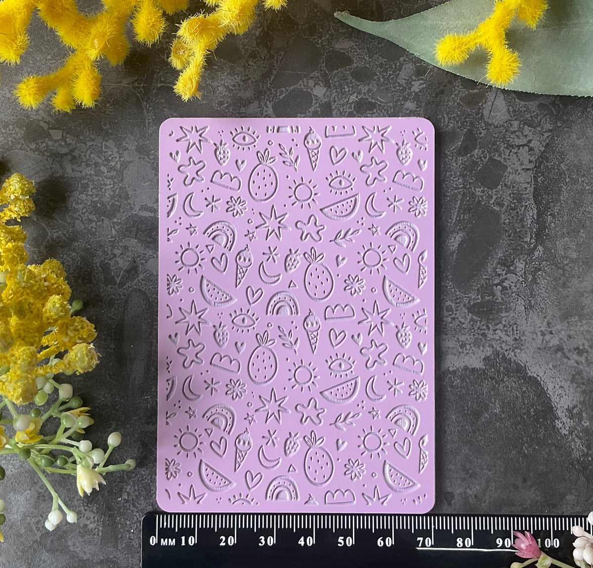 A lavender purple silicone stamp mold with embossed summer-themed designs like ice cream, suns and rainbows, and flowers, displayed on a dark gray textured surface with a black ruler for scale and yellow flowers in the corners.