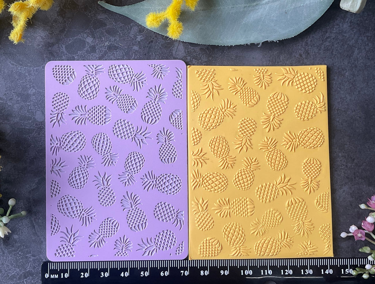 Two rectangular paper sheets with embossed pineapple patterns, one lavender purple and one golden yellow, displayed on a dark gray surface with a black measuring ruler below and yellow flowers and green leaves around the edges.