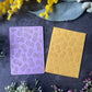 Two rectangular cards or envelopes with pineapple patterns, one in lavender purple and one in mustard yellow, arranged on a dark textured surface with decorative flowers.
