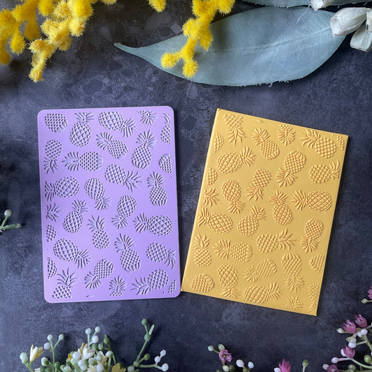 Two rectangular cards or envelopes with pineapple patterns, one in lavender purple and one in mustard yellow, arranged on a dark textured surface with decorative flowers.