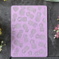 A lavender rectangular stamping or cutting template with a repeating pineapple pattern, displayed on a dark gray textured surface with a black ruler underneath and yellow and pink flowers around it.