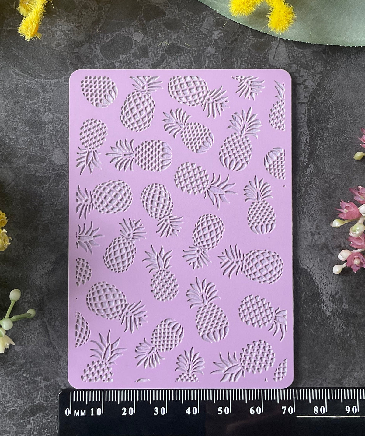 A lavender rectangular stamping or cutting template with a repeating pineapple pattern, displayed on a dark gray textured surface with a black ruler underneath and yellow and pink flowers around it.