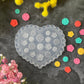 A heart-shaped clear plastic mold with circular floral patterns, surrounded by small colorful flower-shaped embellishments and arranged with yellow and pink flowers on a dark textured background.