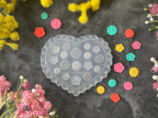 A heart-shaped clear plastic mold with circular floral patterns, surrounded by small colorful flower-shaped embellishments and arranged with yellow and pink flowers on a dark textured background.
