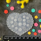 A heart-shaped clear plastic mold or stamp plate with embossed floral and geometric patterns, displayed on a dark textured surface with a black ruler at the bottom and colorful flower buds arranged around it.