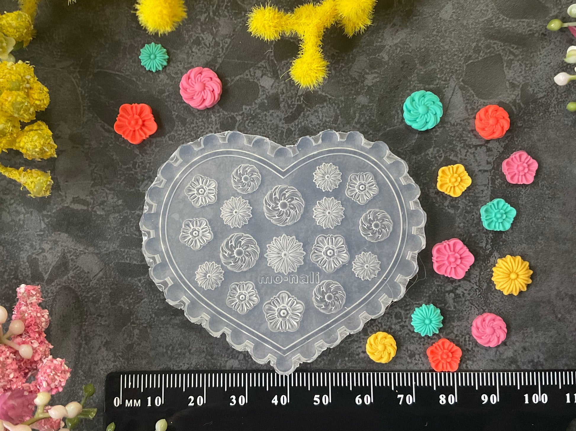 A heart-shaped clear plastic mold or stamp plate with embossed floral and geometric patterns, displayed on a dark textured surface with a black ruler at the bottom and colorful flower buds arranged around it.