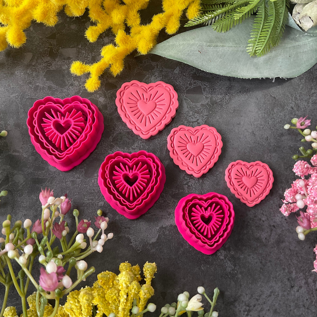 Heart-shaped polymer clay cutters in shades of hot pink, magenta, and coral pink, arranged on a dark gray textured surface surrounded by decorative yellow and pink flowers. Sunburst Heart Beams Polymer Clay Cutter