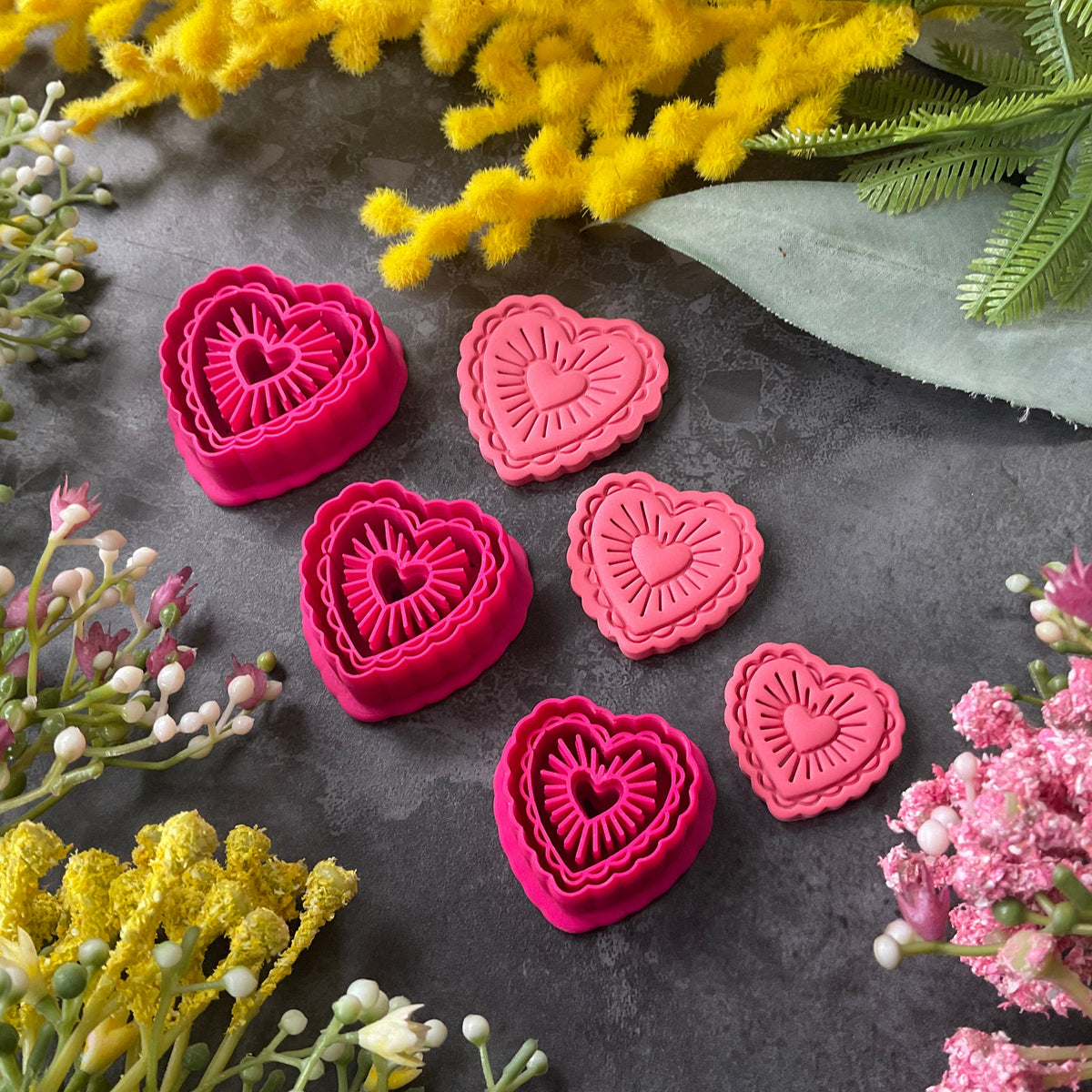 Heart-shaped polymer clay cutters in shades of hot pink, magenta, and coral pink, arranged on a dark gray textured surface surrounded by decorative yellow and pink flowers. Sunburst Heart Beams Polymer Clay Cutter