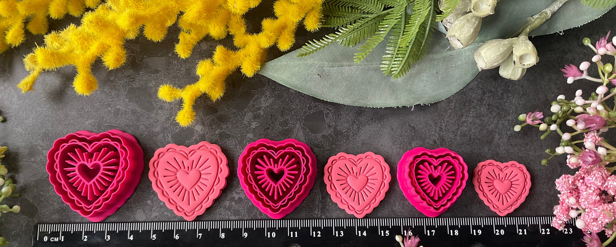 Heart-shaped polymer clay cutters in shades of hot pink, magenta, and coral pink, arranged on a dark gray textured surface surrounded by decorative yellow and pink flowers. Sunburst Heart Beams Polymer Clay Cutter
