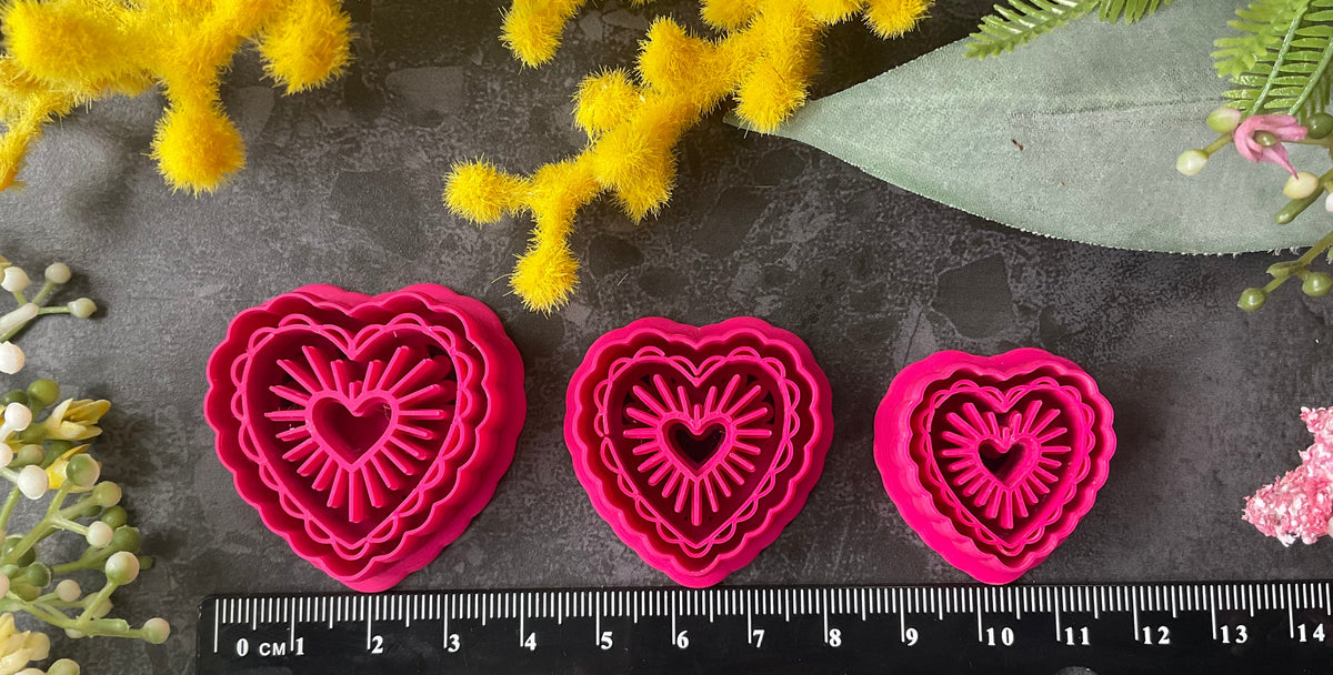 Heart-shaped polymer clay cutters in shades of hot pink, magenta, and coral pink, arranged on a dark gray textured surface surrounded by decorative yellow and pink flowers. Sunburst Heart Beams Polymer Clay Cutter