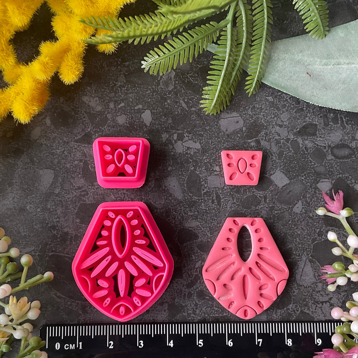 Bright pink3d printed polymer clay cutters in different sizes arranged on a dark textured surface, surrounded by decorative flowers and greenery. Terracotta Tale Polymer Clay Cutter Set