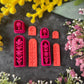 A set of 3d printed flower-shaped polymer clay cutters in different sizes arranged on a dark textured surface with a black ruler below and yellow and pink flowers around the edges. The cutters are hot pink and orange, Winter Meadow Polymer Clay Cutter Set.